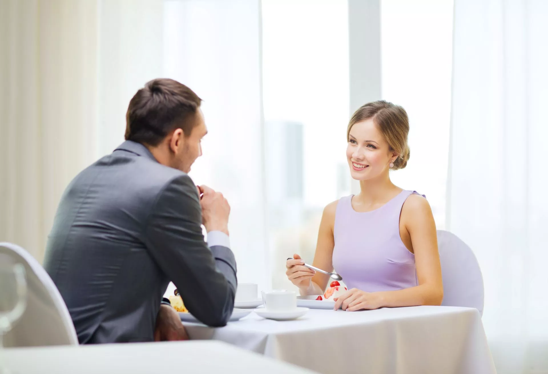 一名女子在明亮室內餐廳座位上微笑用餐，手持湯匙享用甜點，對面男子身穿西裝傾身交談，桌上擺放白色餐具，呈現輕鬆愉快的約會用餐情境。